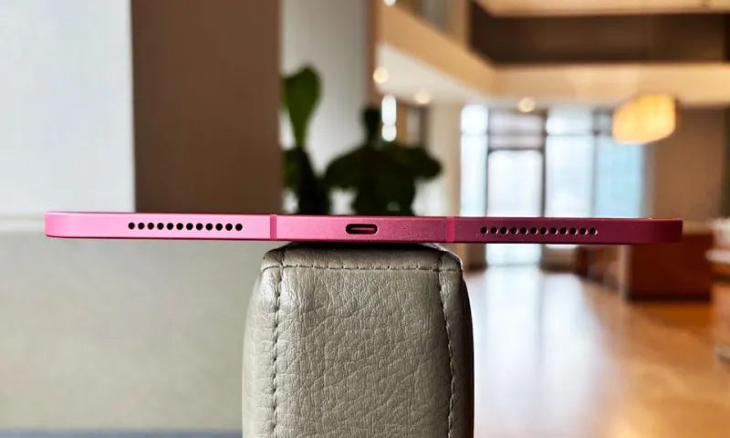 A pink Apple iPad (A16) rests on the flat arm rest of a light brown leather chair, with its bottom edge facing the camera, showing off the device's thin profile along with its speaker grilles and USB-C port.
