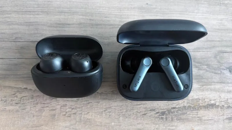 Two black pairs of wireless earbuds, the JLab Go Pop ANC and EarFun Air Pro 4, rest on a light brown wooden table with their cases open, exposing the earbuds as they charge inside.